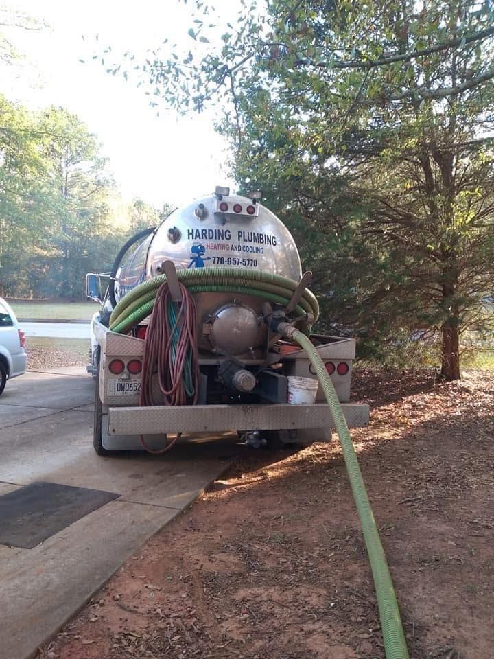 A plumbing truck with a large tank and green hose parked on a driveway in front of trees.