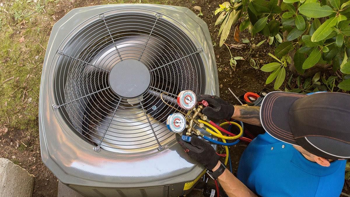 Person in blue shirt working on an AC unit, connecting gauges to check the pressure.