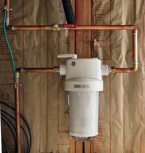 Copper pipes connected to a white water filter. Red valve on upper pipe. Green wire grounded to copper.
