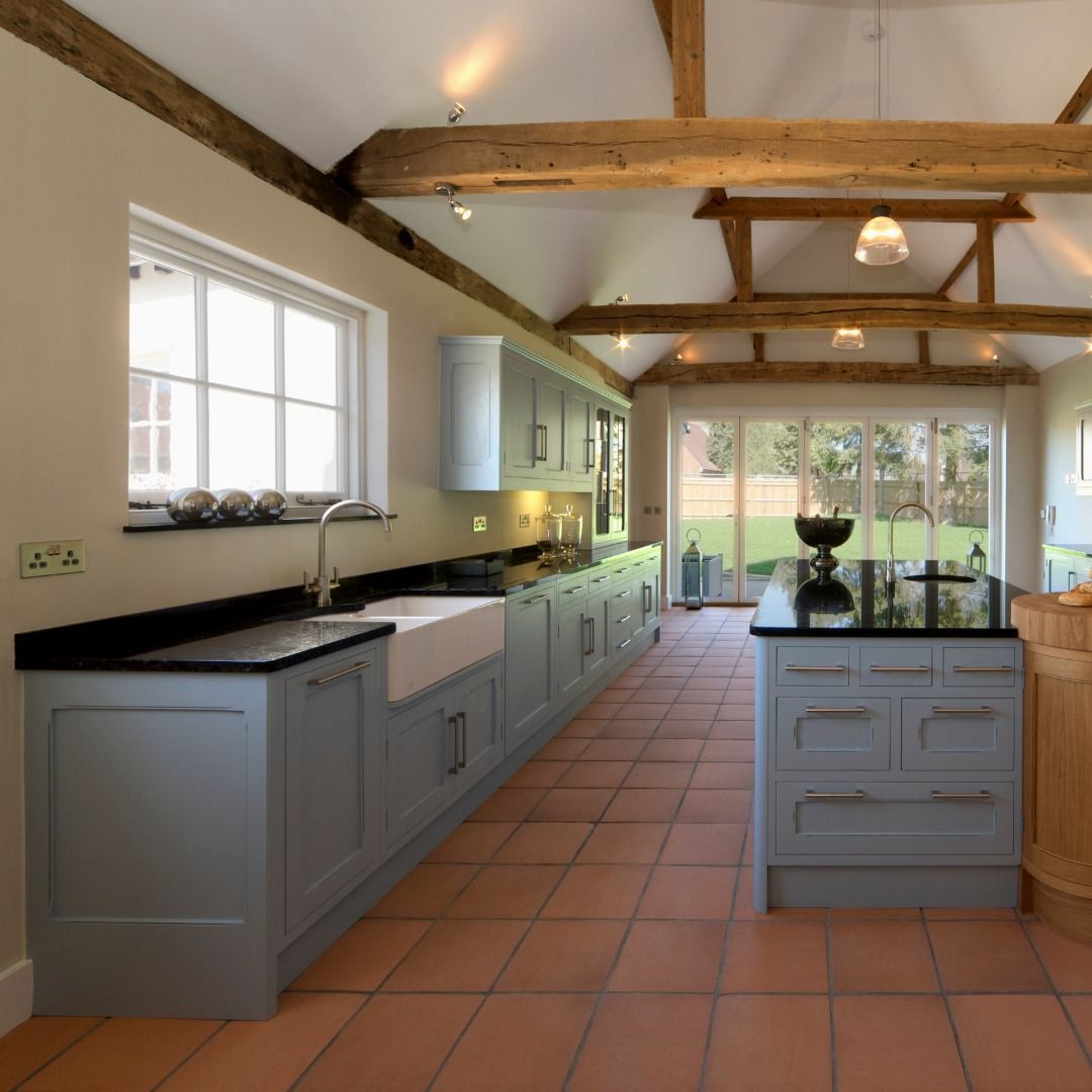 Spacious kitchen with gray cabinets, black countertops, and terracotta tile floor, with exposed wood beams and large windows.