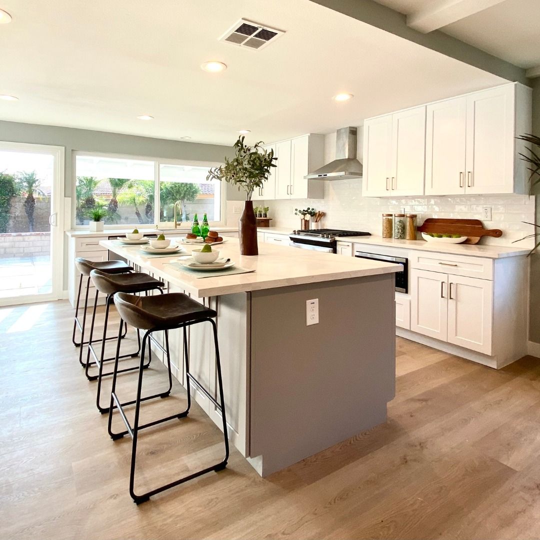 Bright kitchen with white cabinets, gray island, and light wood flooring. Black bar stools are at the island.