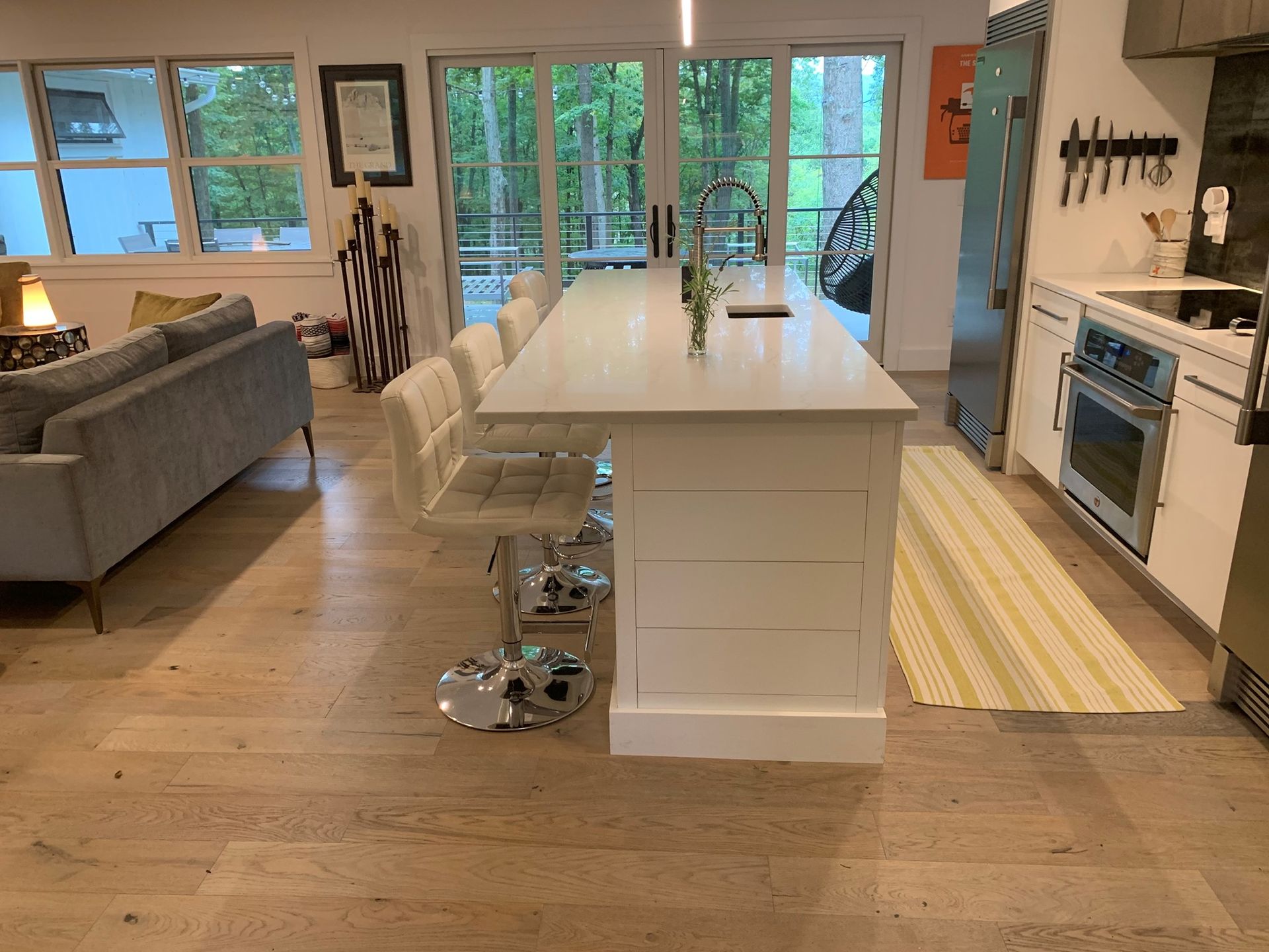 Modern kitchen with island, white cabinets, and wood flooring. Large windows look out to trees.