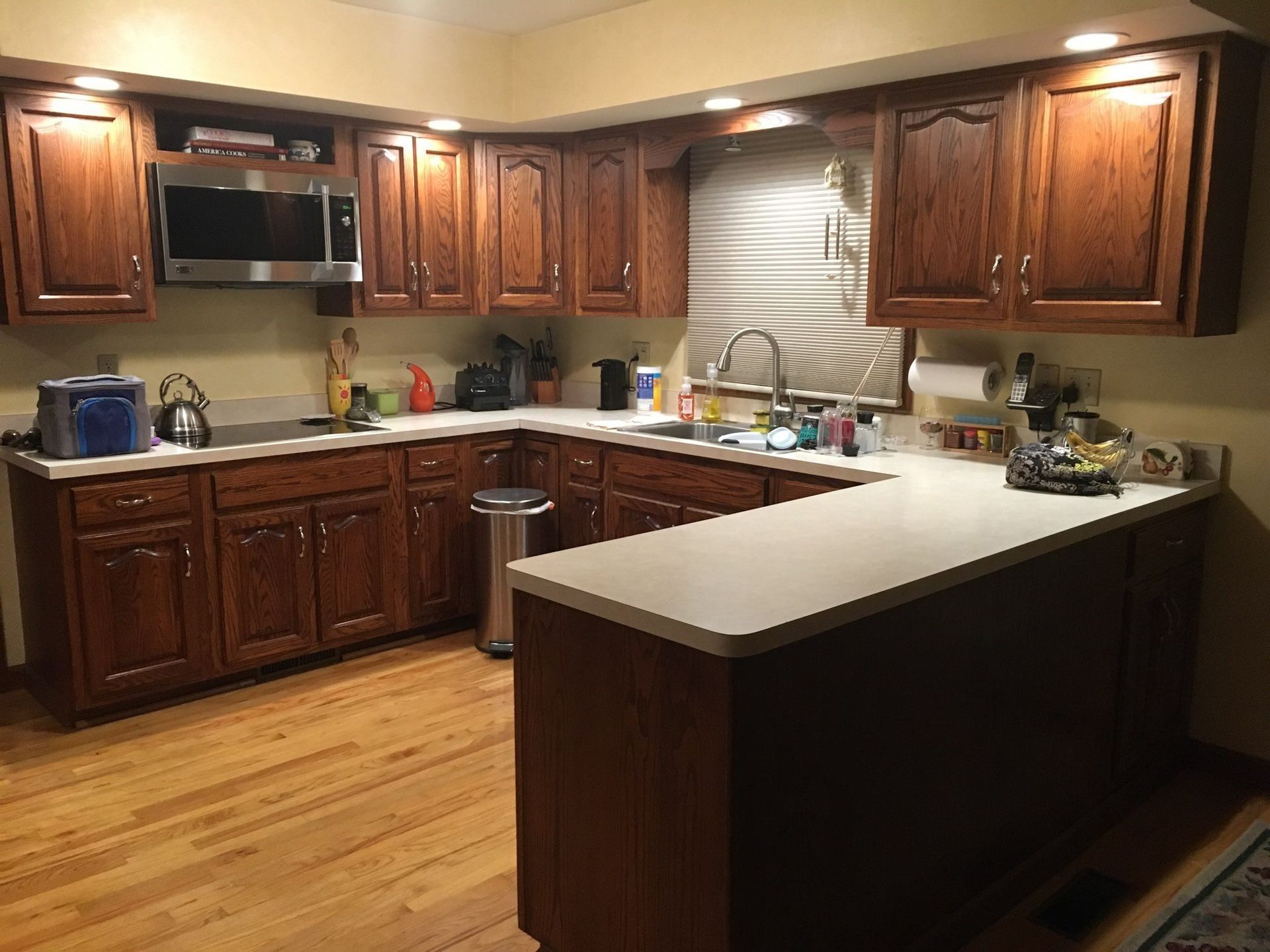 A kitchen with brown cabinets, light countertops, and wooden floors. A microwave and sink are visible.
