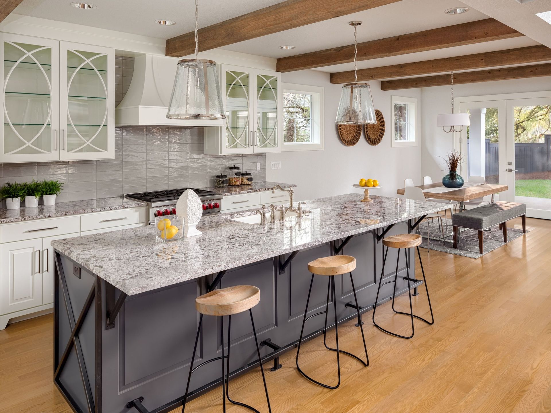 Modern kitchen with gray island, granite countertop, light wood floors, and wooden beams.