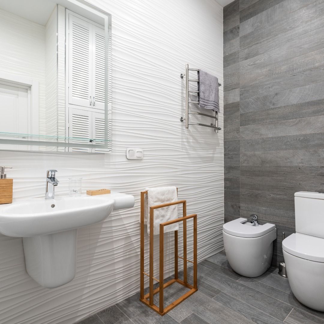 Modern bathroom with white sink, toilet, and textured walls; gray tile accent wall; wooden towel rack.