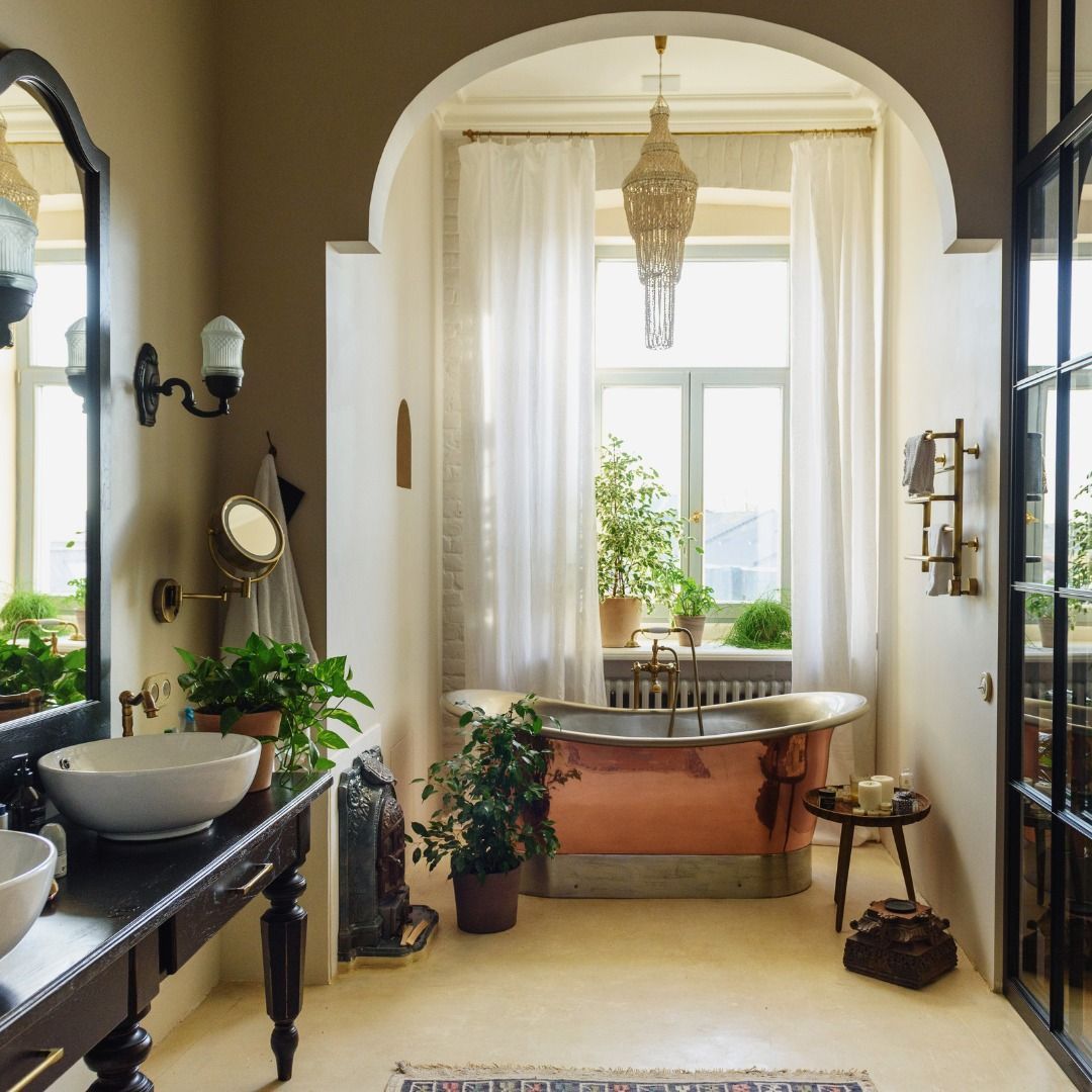 Elegant bathroom with copper tub, arched window, and plants; neutral tones and vintage style.