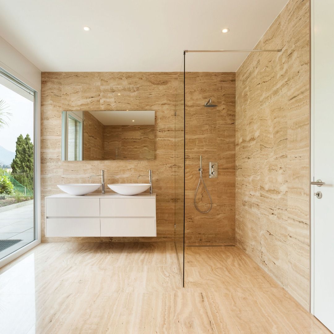 Modern bathroom with travertine walls and floors, glass shower, white vanity, and large window.