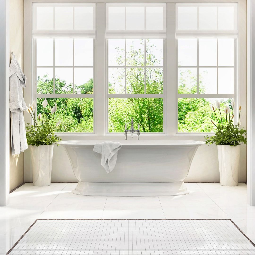 A bright bathroom with a white tub in front of three large windows overlooking lush greenery.