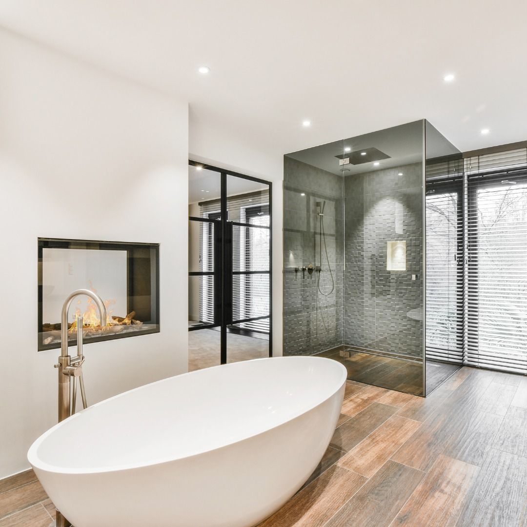 Modern bathroom with white tub, fireplace, glass shower, wooden floor, and windows.