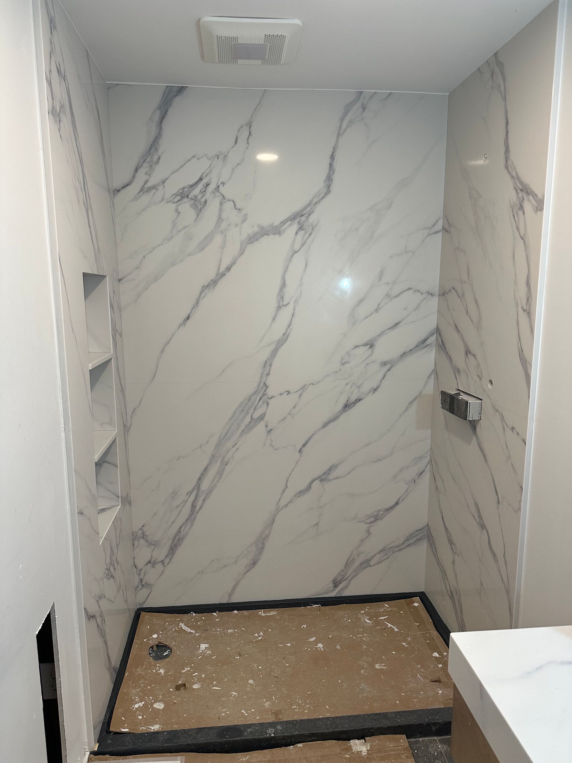 Marble-look tiled shower with built-in shelves. Construction incomplete, gray floor, white and gray veined tiles.