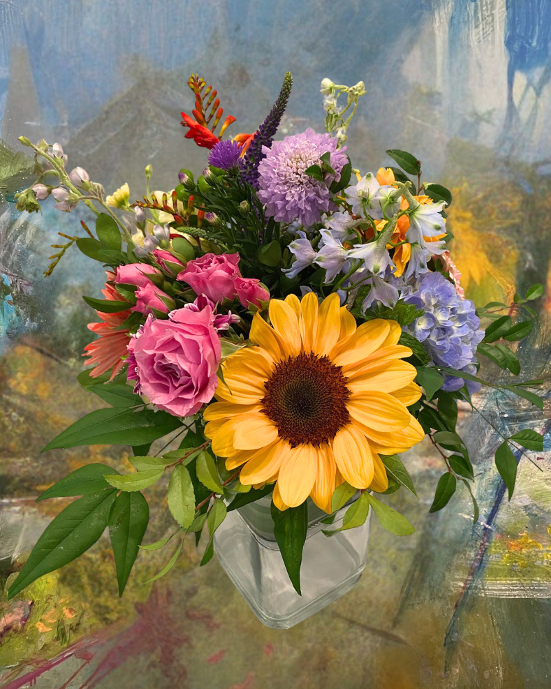 Colorful bouquet in a white vase with a large yellow sunflower, pink roses, and purple blossoms