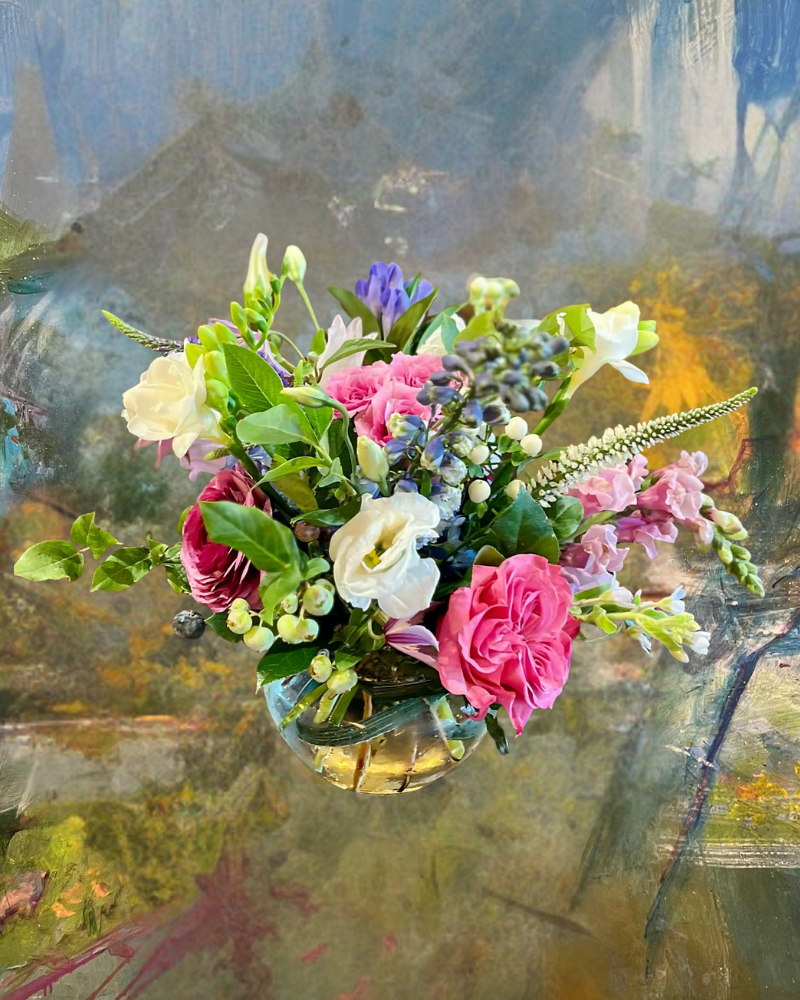 Colorful bouquet of pink, white, yellow, and purple flowers in a glass vase against a muted abstract background