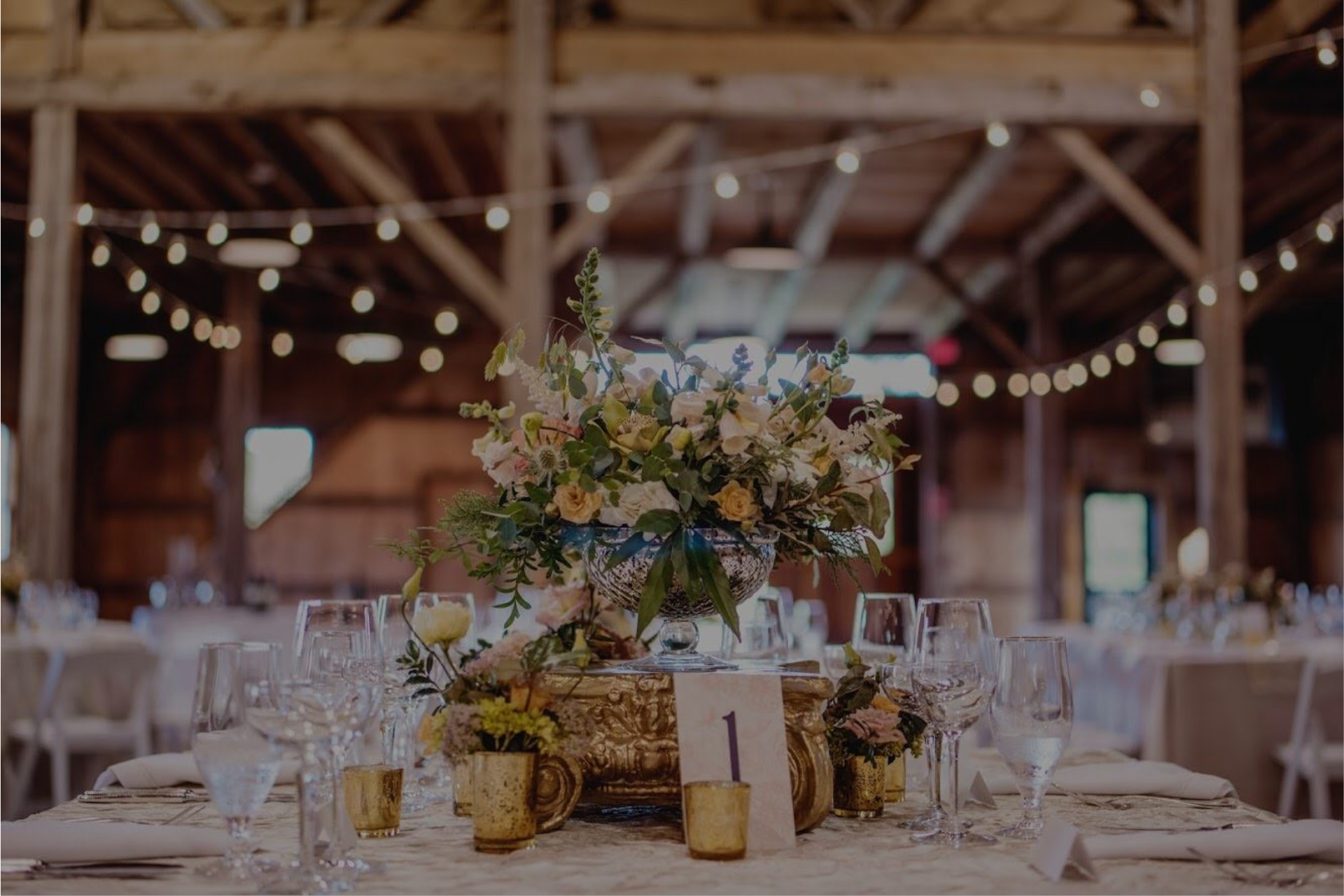 Rustic reception table with floral centerpiece, candles, and glassware under string lights in a barn