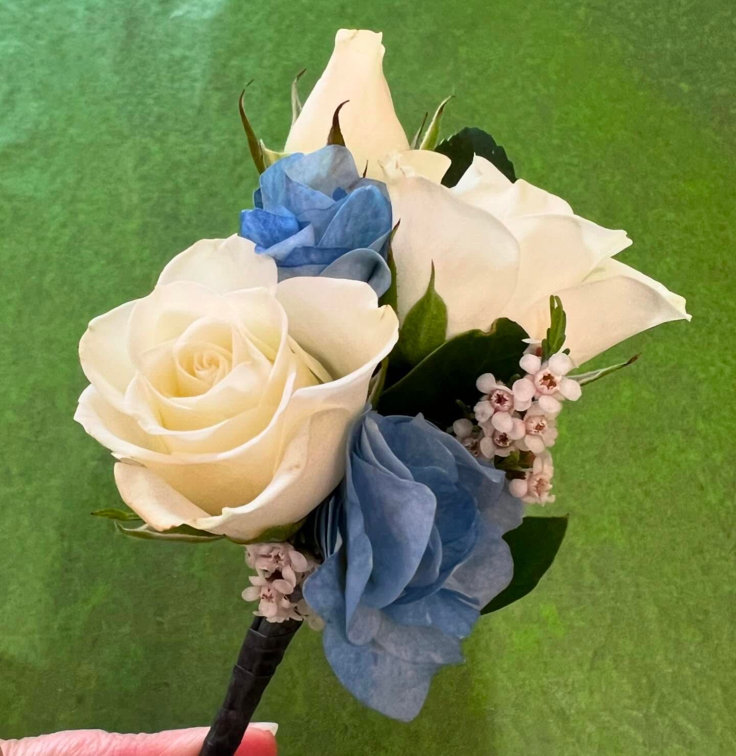 White and blue rose boutonniere with small pink flowers and green leaves, held by a black stem against a green background.