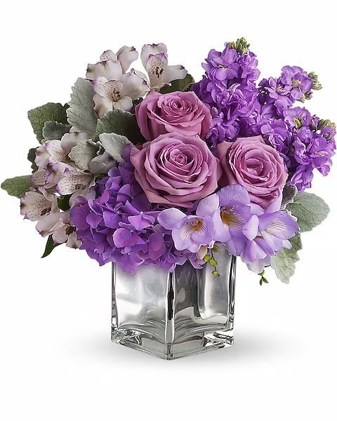 Purple and lavender floral arrangement in a square glass vase. Roses, stock flowers, and other blooms.