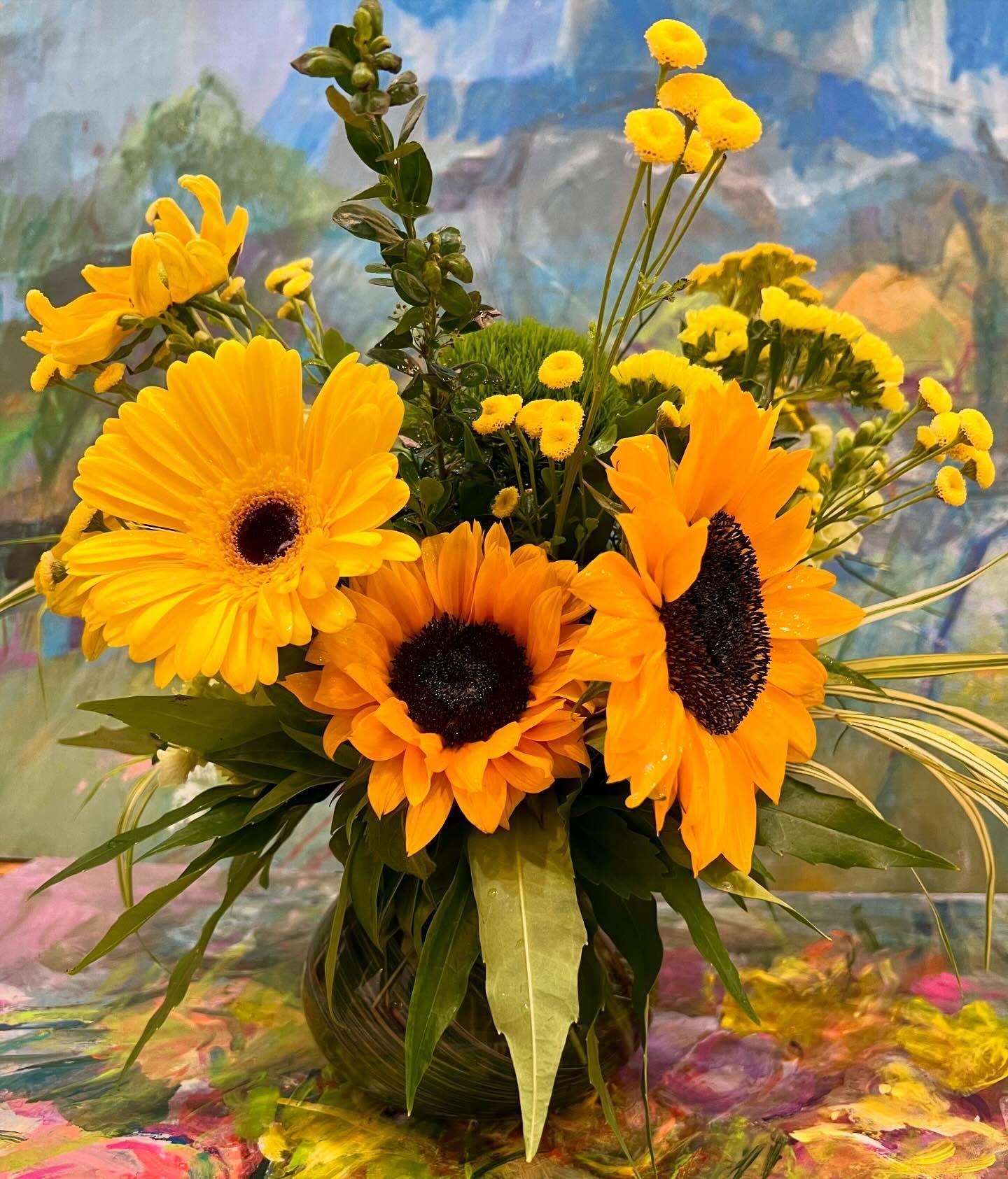 Vibrant yellow sunflowers and gerbera daisies in a glass vase, against a colorful painted backdrop.