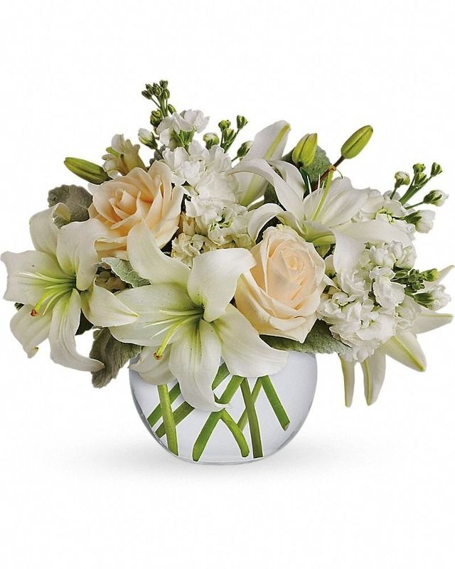 A floral arrangement of pale peach roses and white lilies with green stems in a clear, round glass vase.