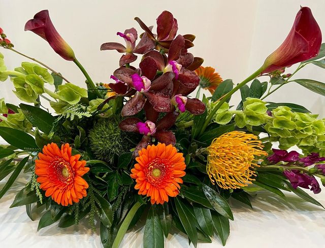 Floral arrangement with orange gerbera daisies, burgundy orchids, and green accents.
