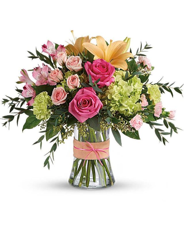 A floral bouquet with pink roses, peach lilies, and green hydrangea in a clear glass vase wrapped with a burlap ribbon.