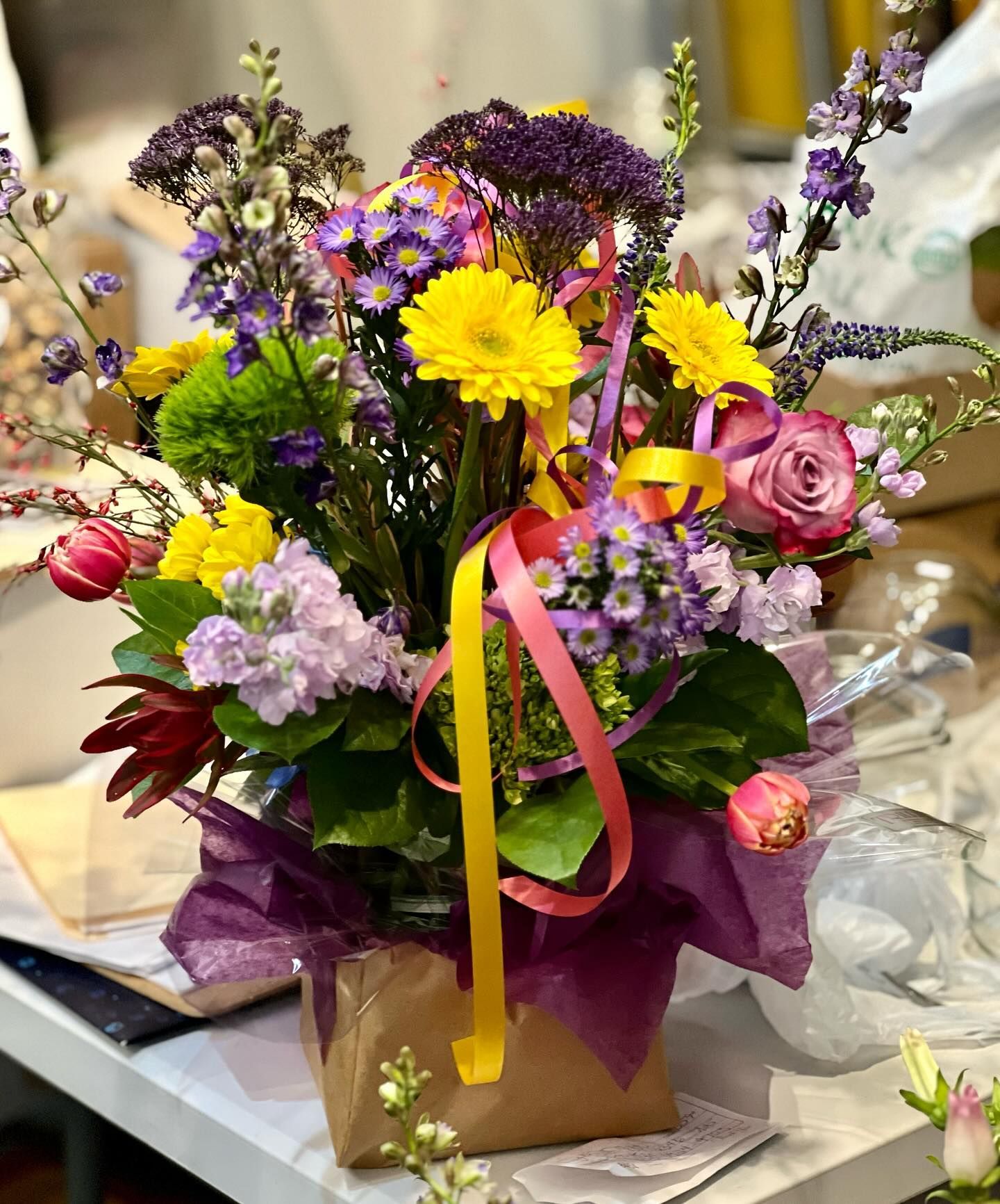 Colorful flower arrangement in a brown paper-wrapped container, with ribbons and varying blooms.