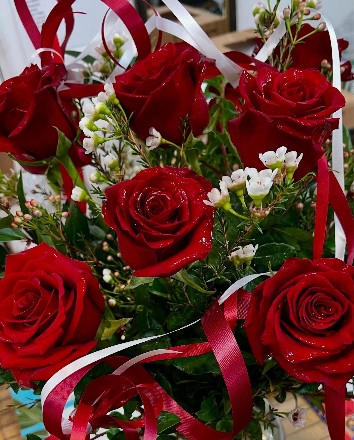 Close-up of a bouquet of six red roses with white and red ribbons and small white flowers.