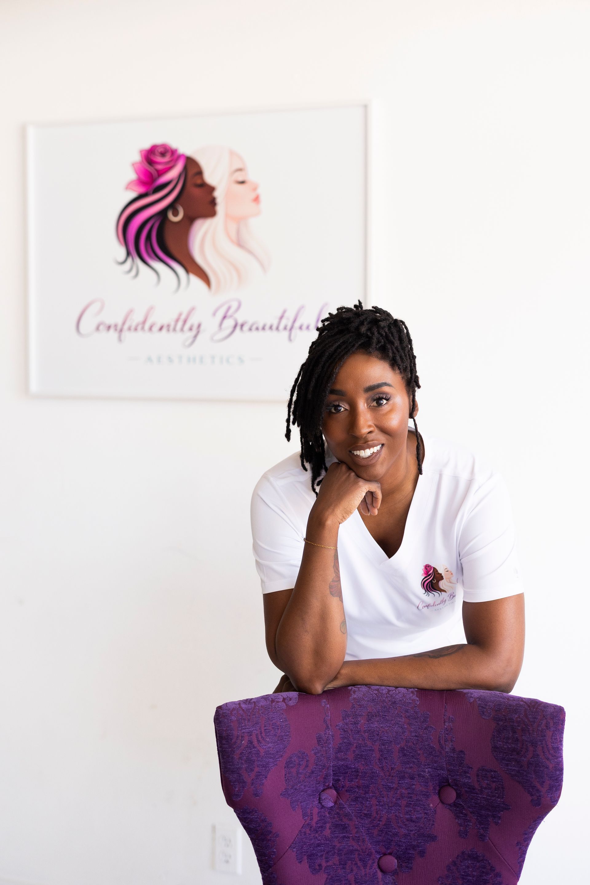 Woman leaning on purple chair in a bright room beside a wall logo for “Confidently Beautiful.”