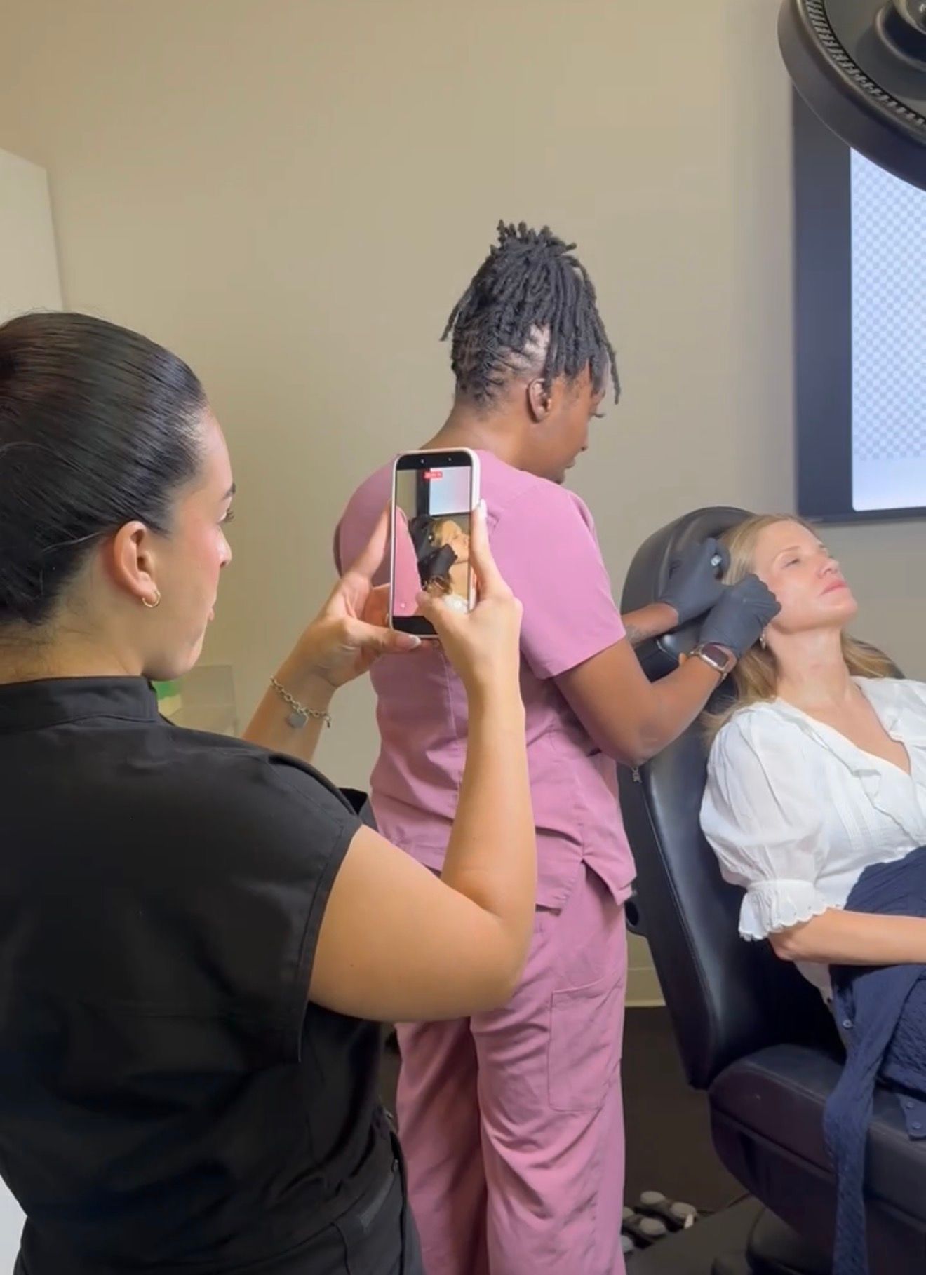 A person giving a facial treatment while another records it on a phone, with a client reclining in a salon chair.