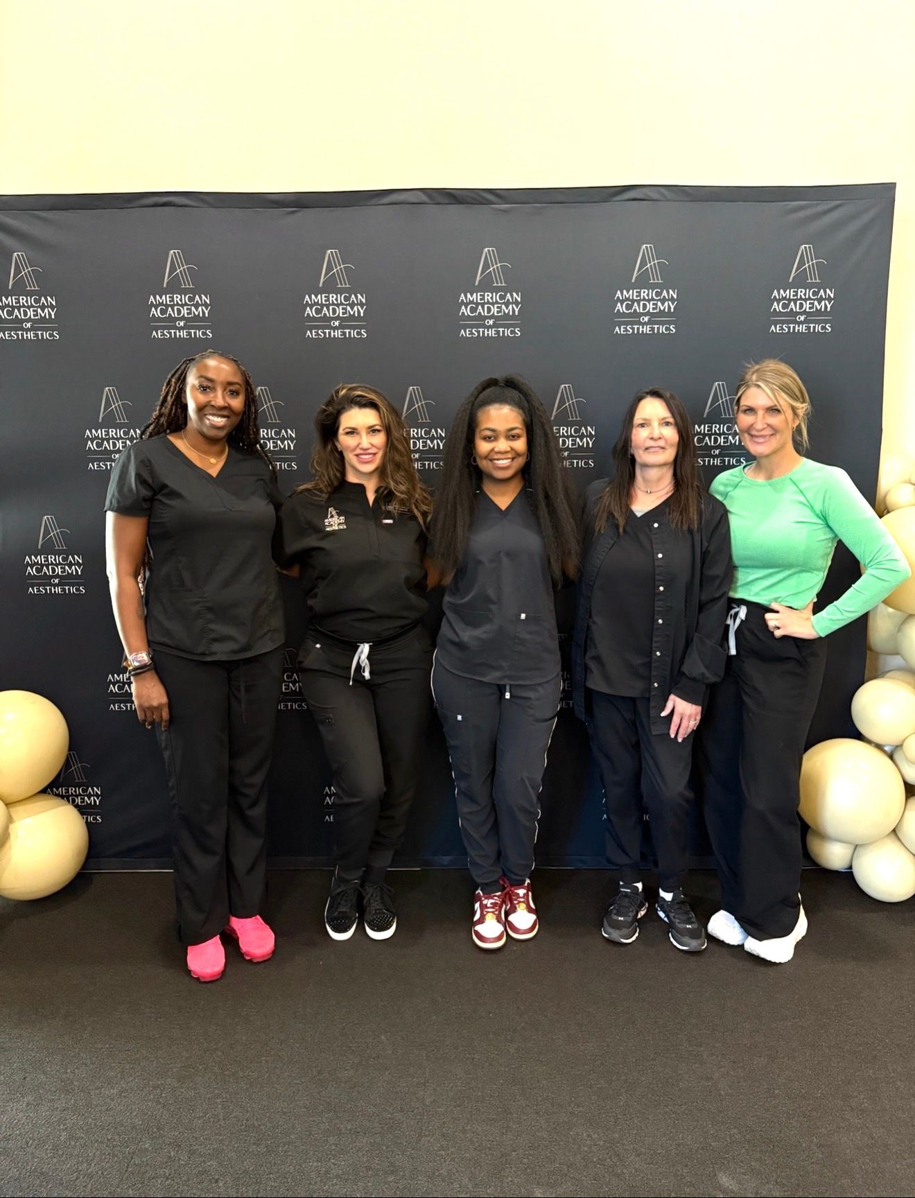 Five women pose in front of a branded backdrop with gold balloons at an event.