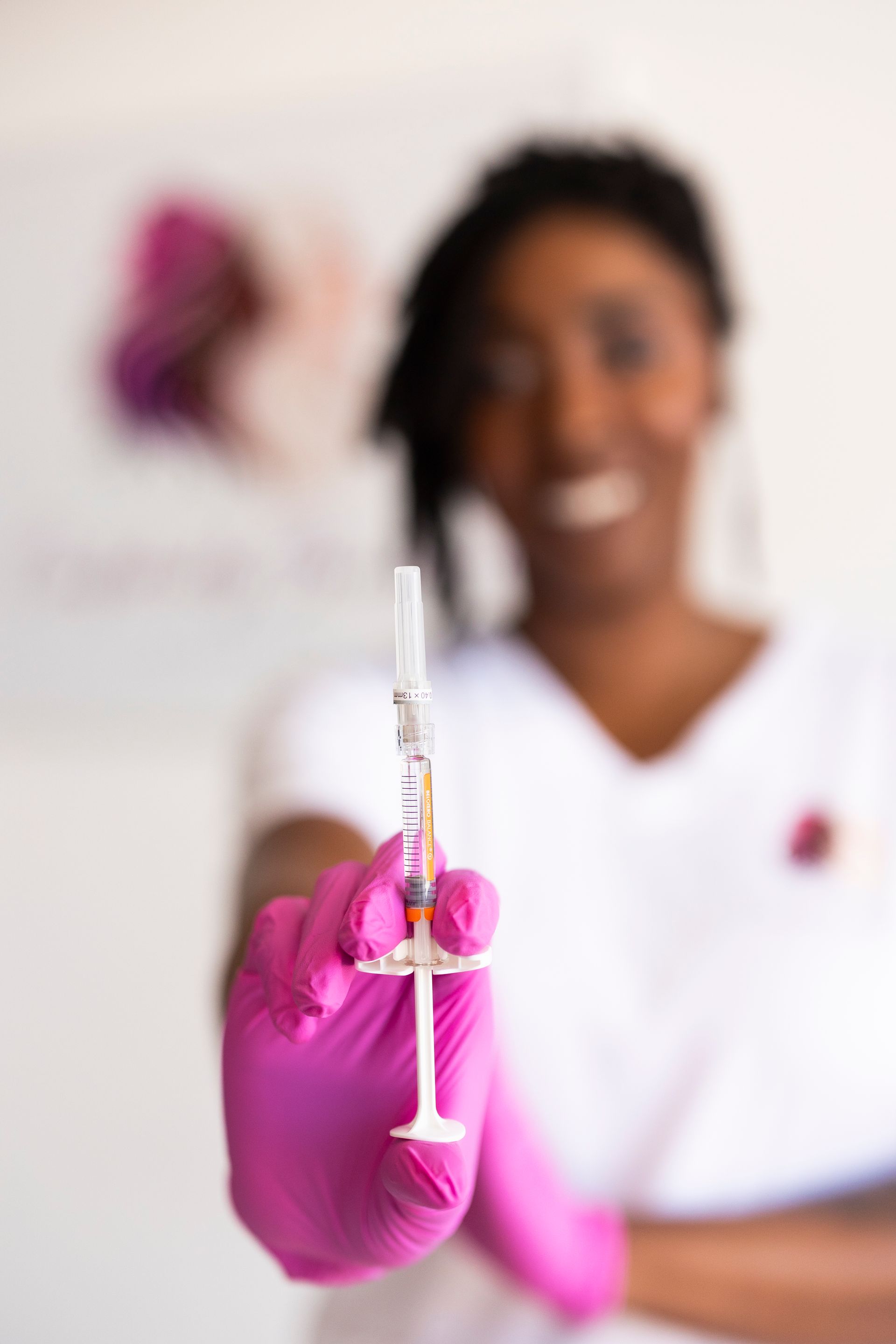 Gloved hand holding a syringe toward the camera, with a smiling clinician blurred in the background.