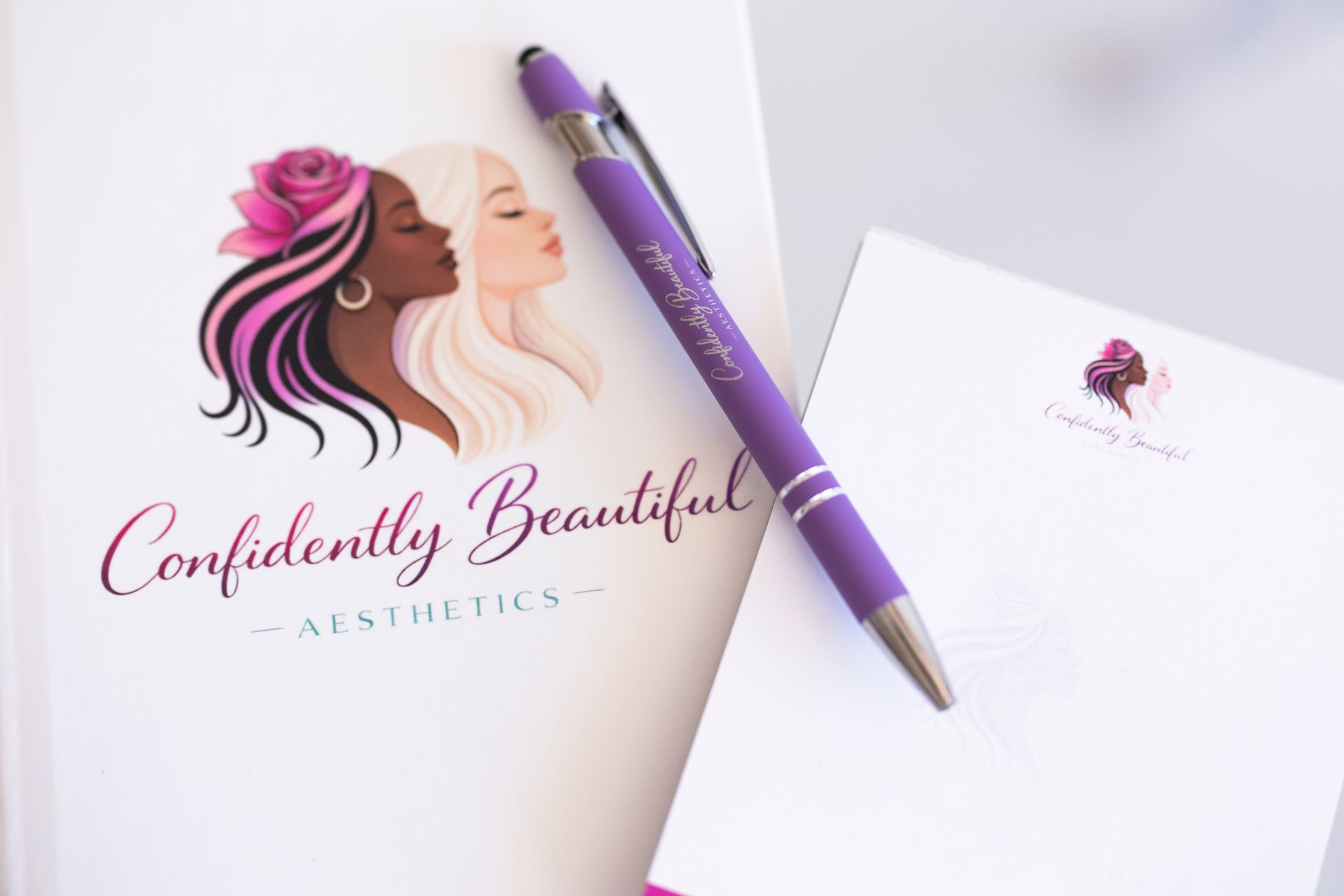 Confidently Beautiful Aesthetics logo with pink and purple makeup faces and pen on white stationery