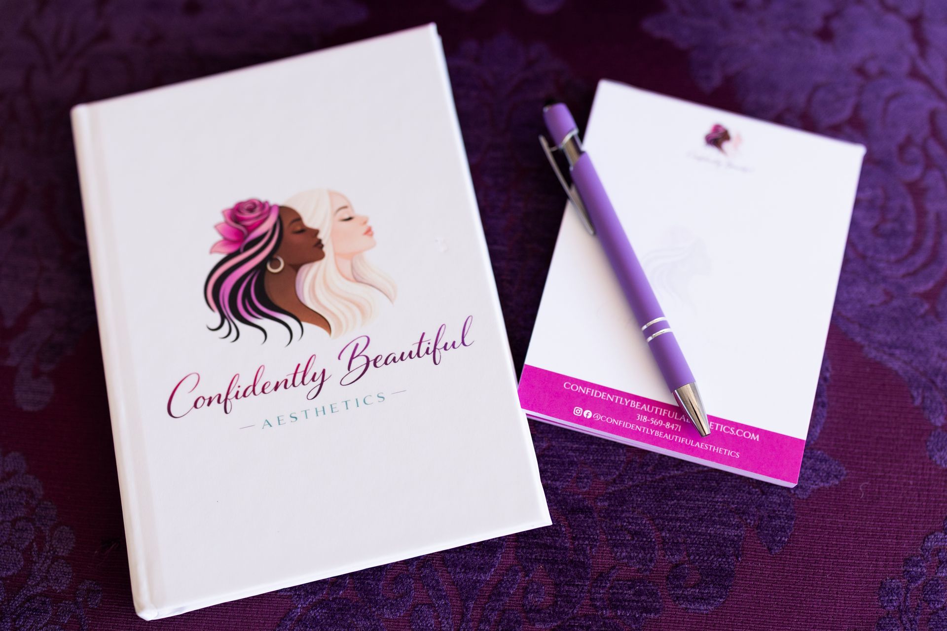 White notebook and purple pen on a purple textured surface, with a fashion logo and pink accent notebook.