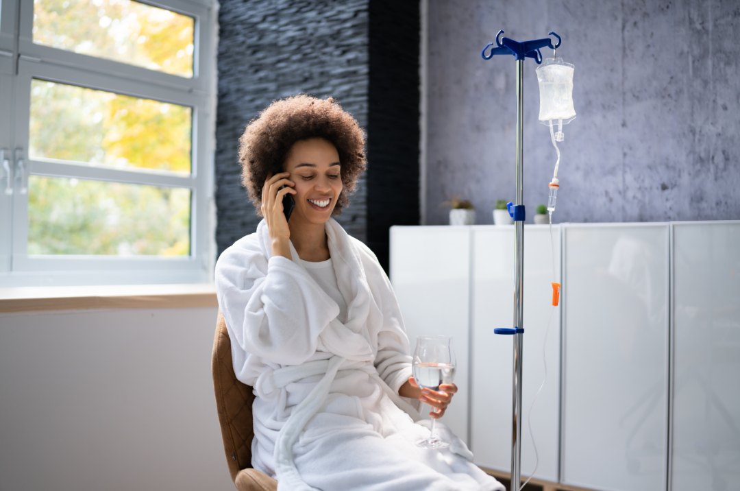 Person in a white robe smiling in a medical room beside an IV stand