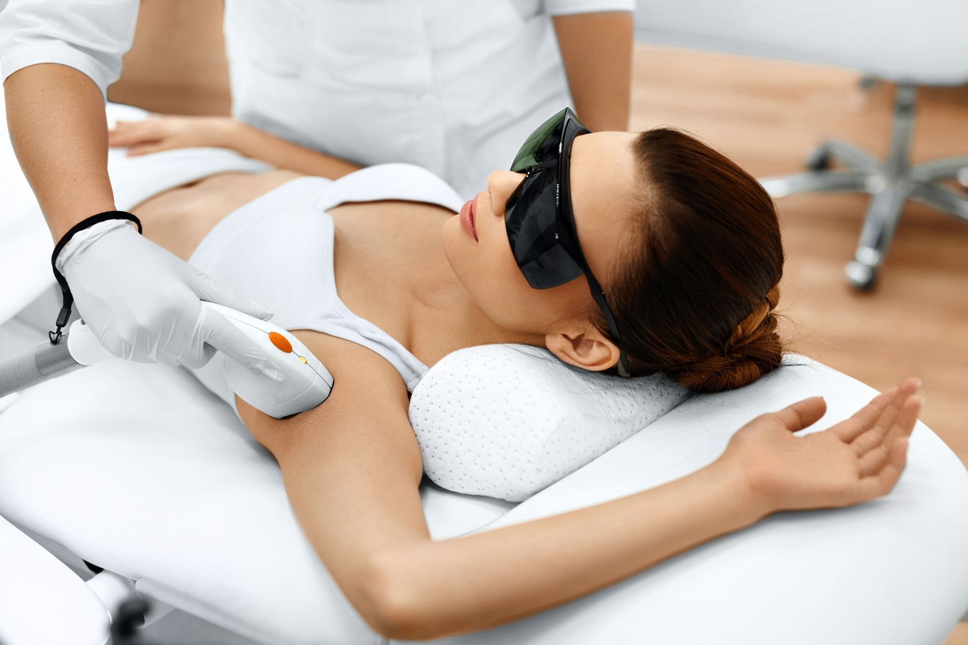 Woman receiving a facial treatment on a spa bed, wearing protective goggles and holding an orange handheld device