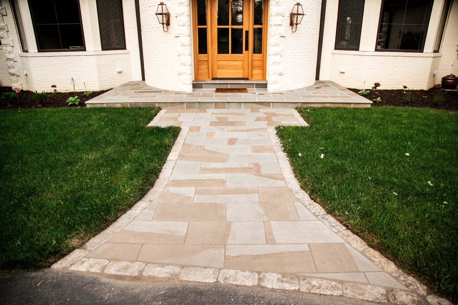 Stone pathway leading to a wooden door, flanked by green grass and a white brick house.