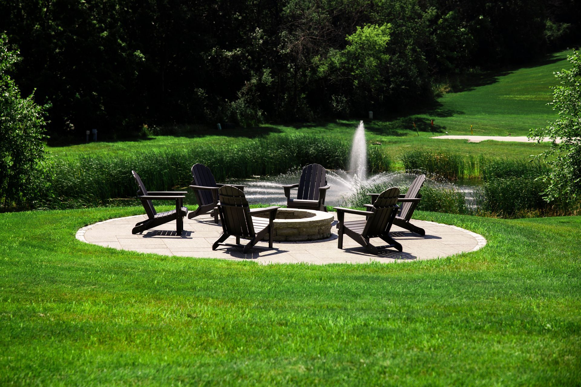 Circle of chairs around a fire pit on a circular patio, in a grassy backyard with a fountain.