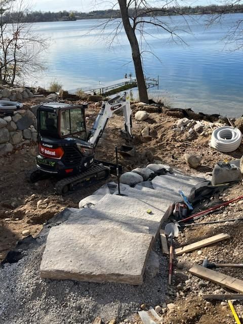 A small excavator is sitting on top of a pile of rocks next to a body of water.