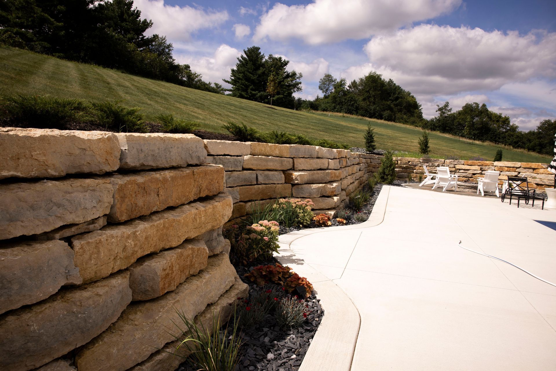 A stone wall surrounds a patio with chairs and a lawn in the background.