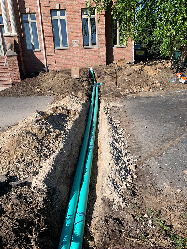 A green pipe is being installed in the dirt in front of a brick building.