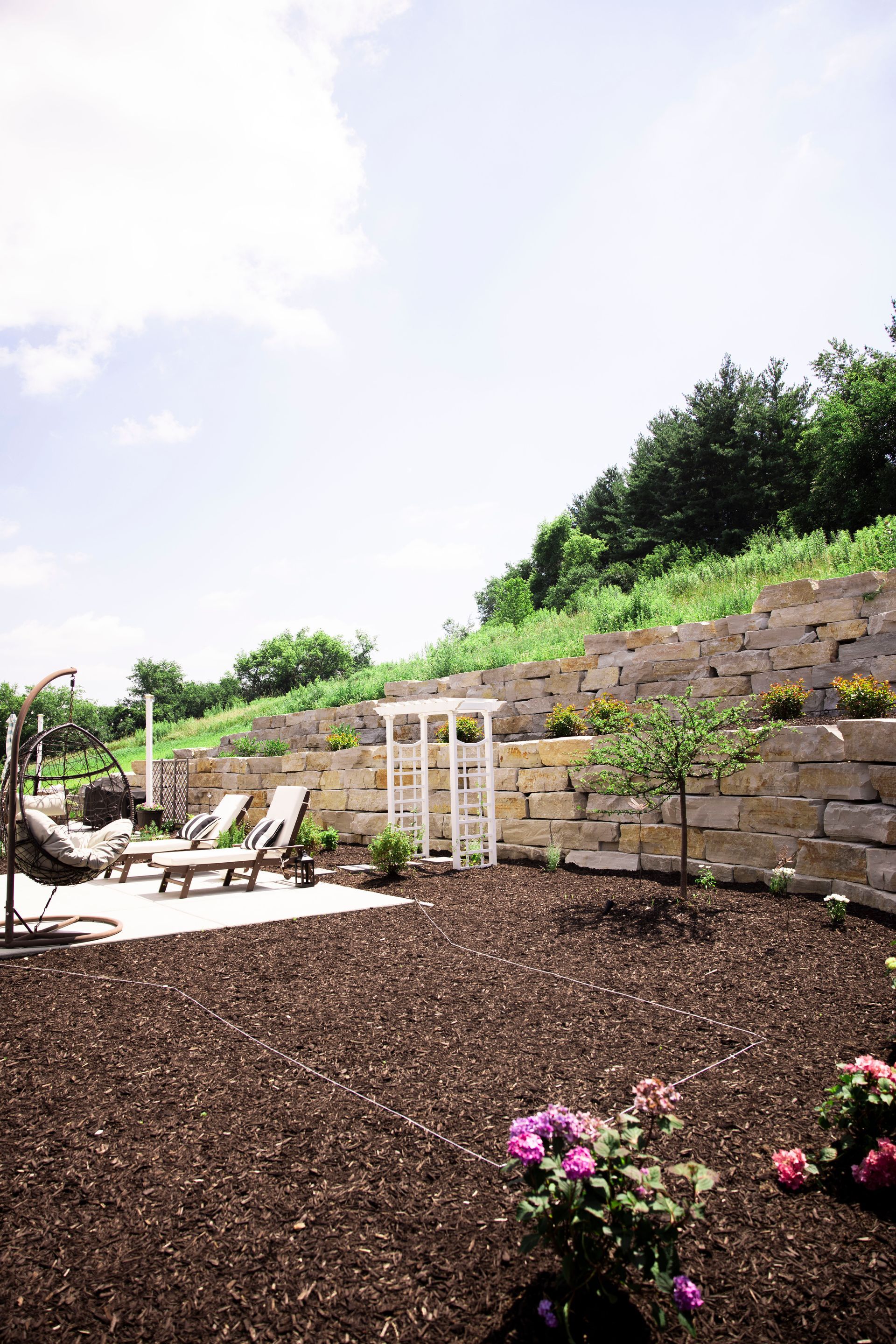 Backyard with stone retaining wall, lounge chairs, and garden bed with mulch and flowers under a blue sky.