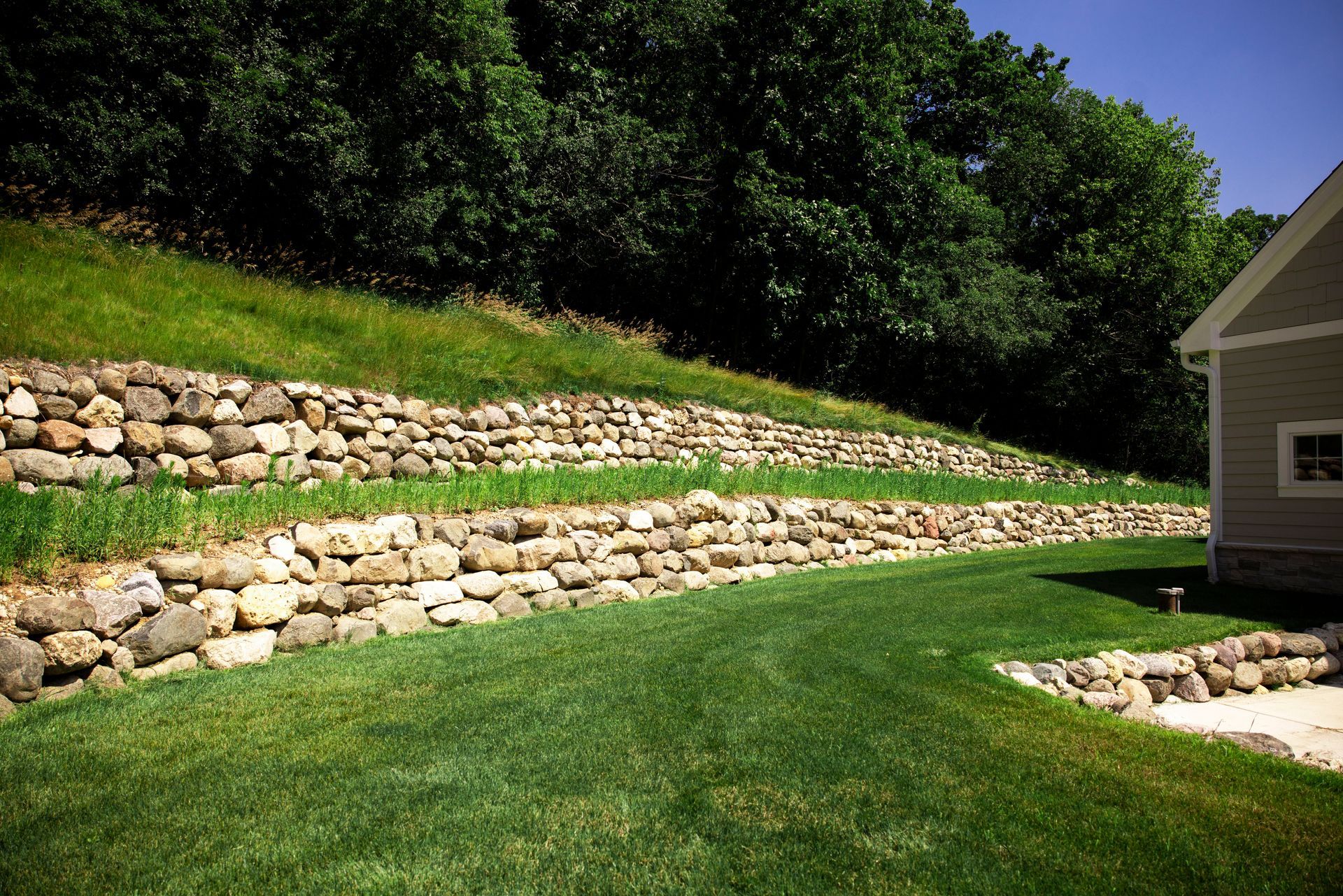 Stone retaining walls on a grassy hillside, next to a house with a green lawn.