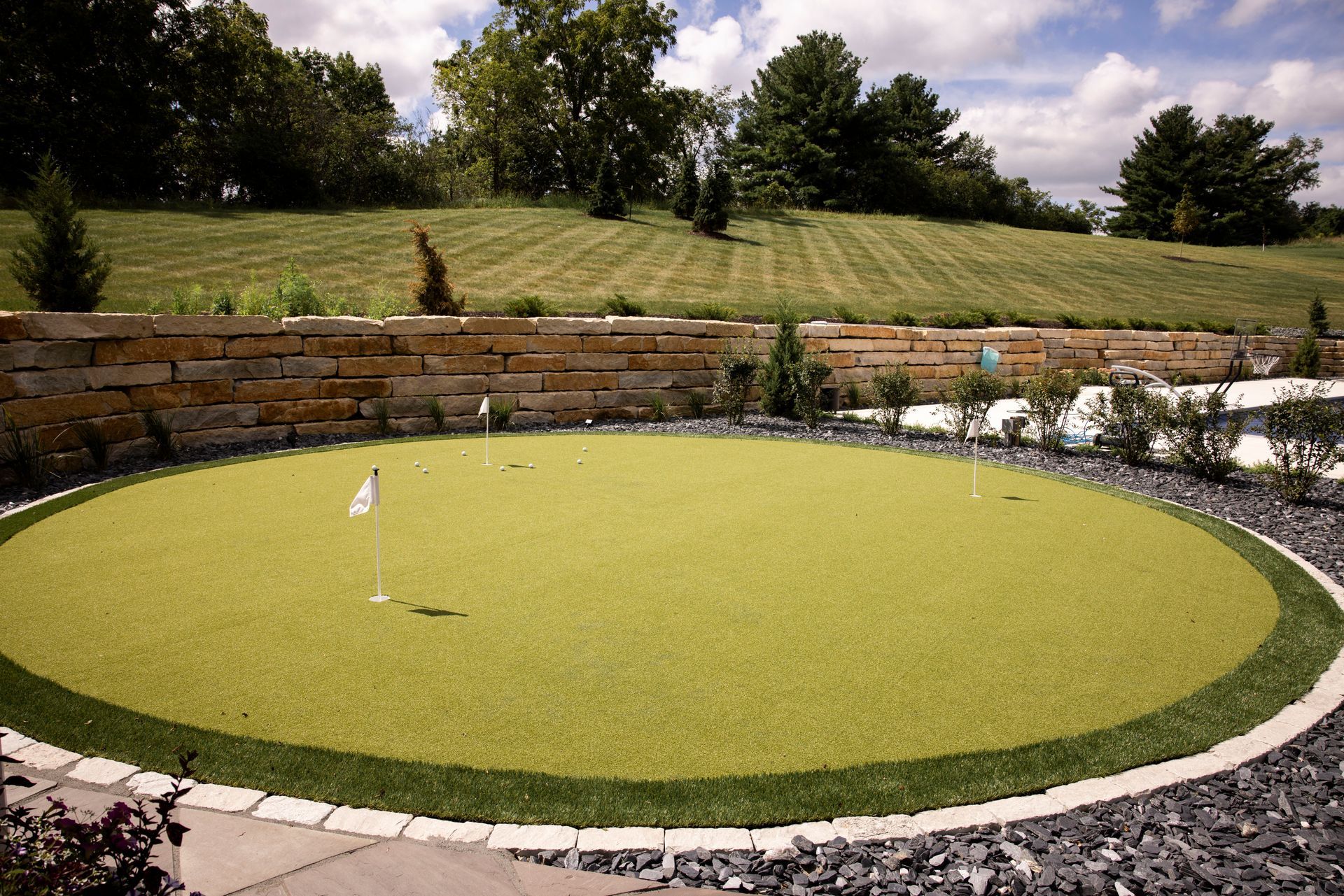 A circular artificial putting green with a flag, surrounded by dark stones and a stone wall, in a yard.