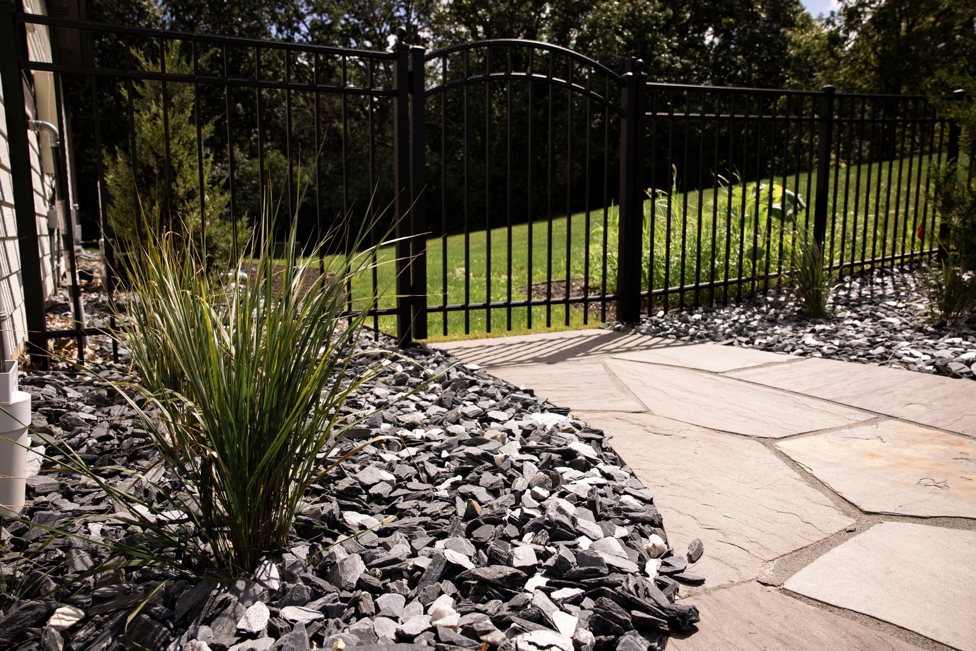 Black iron fence with gate, bordering a stone path and rock bed with green grass visible beyond.