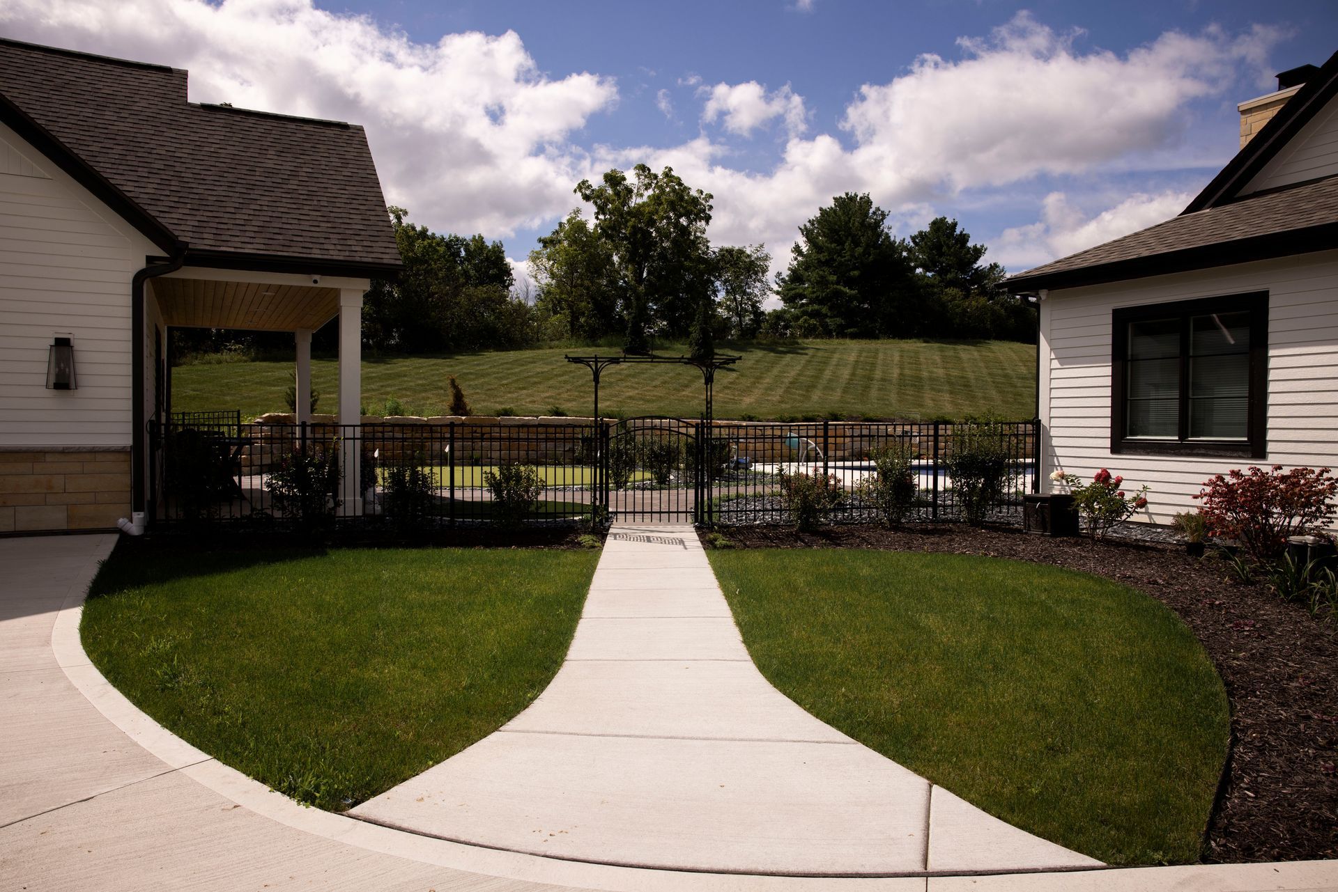 Pathway through green lawn, between two white buildings, leading to a fenced area under a partly cloudy sky.