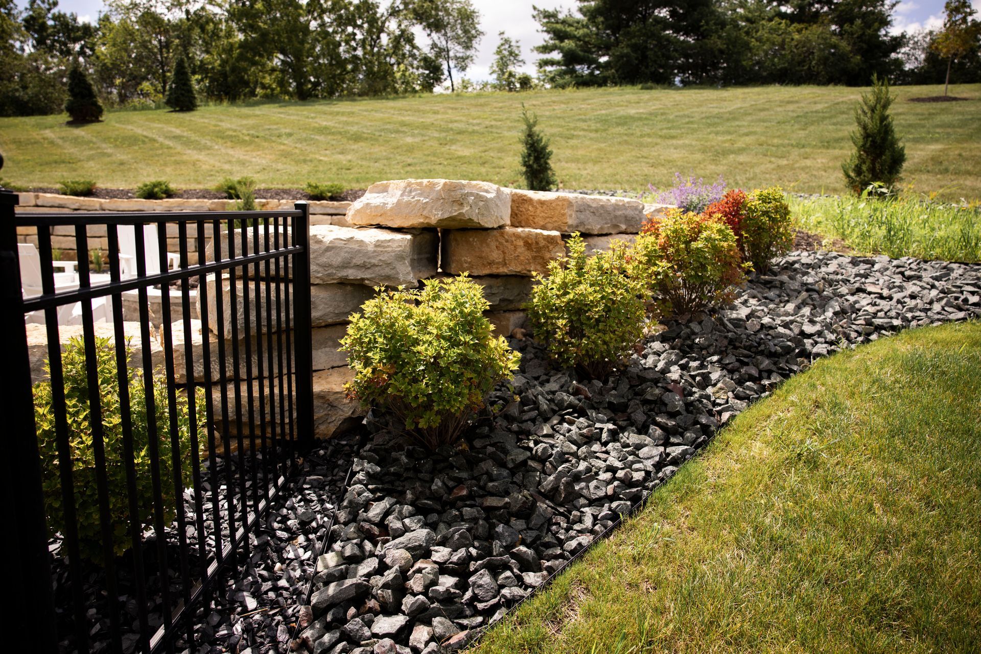 Black fence with stone wall, shrubs, and black rock landscaping along a grassy lawn.