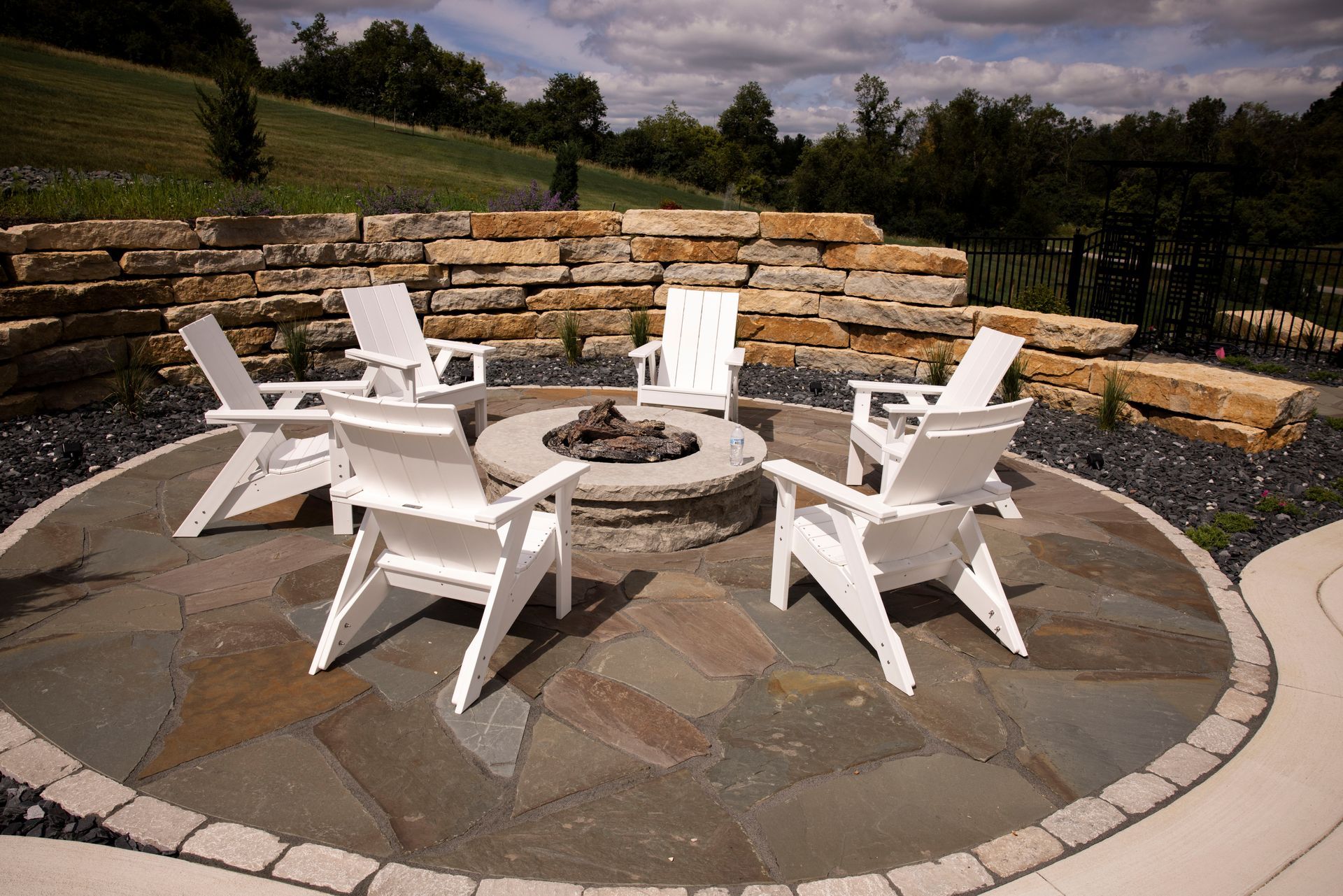 White chairs around a stone fire pit on a circular patio, set against a stone wall and grassy hill.