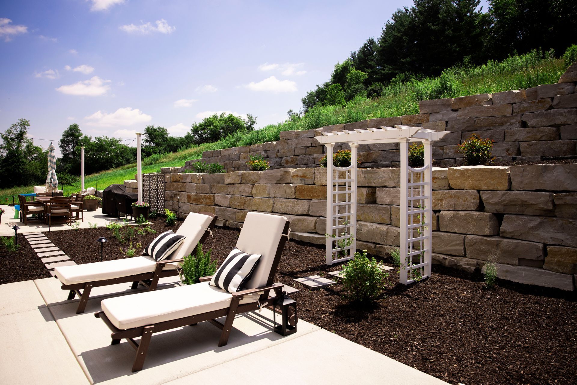 Two lounge chairs on patio, with striped pillows, next to a stone wall and white arbor.