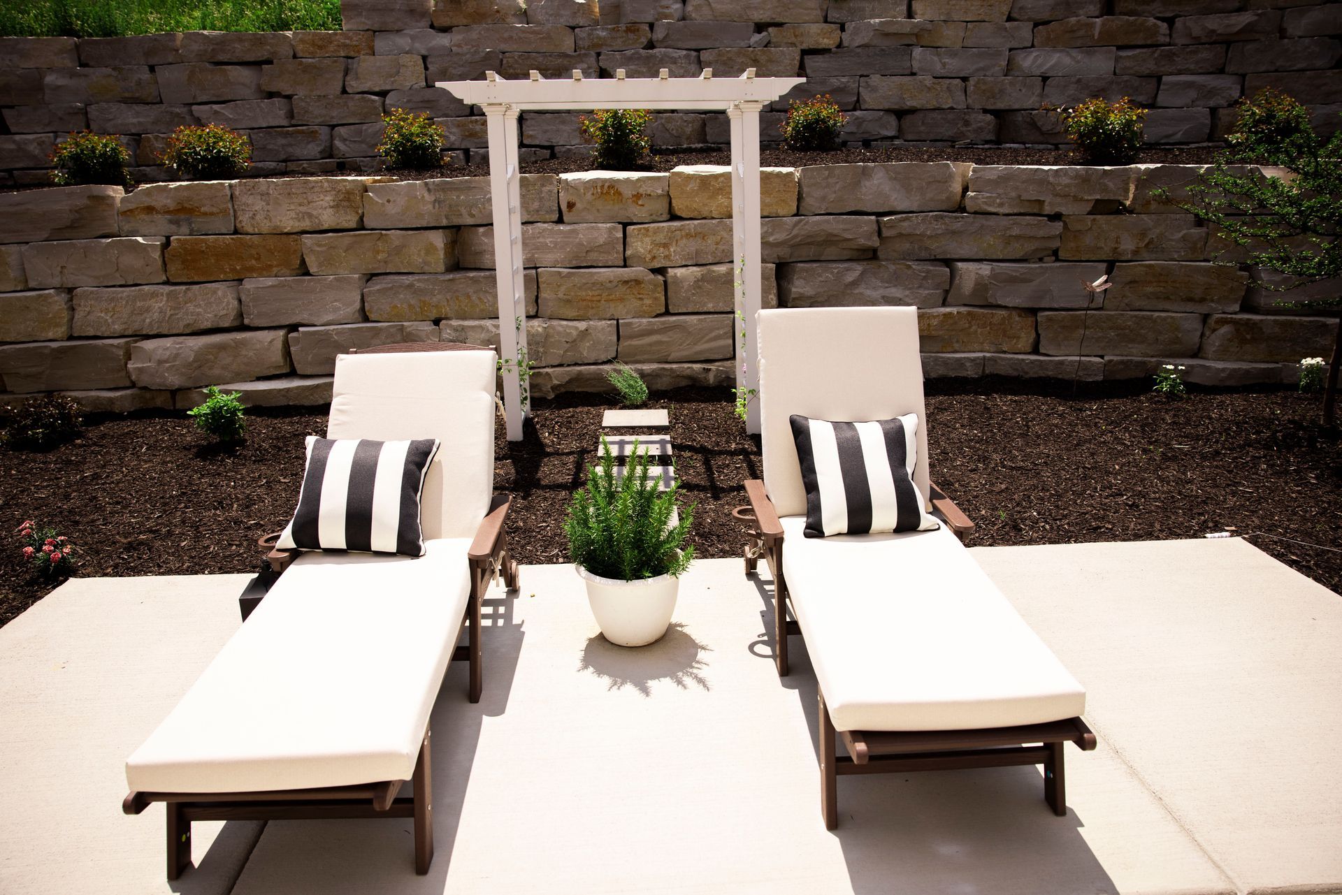 Two lounge chairs with black and white striped pillows under a white trellis, near a stone wall.