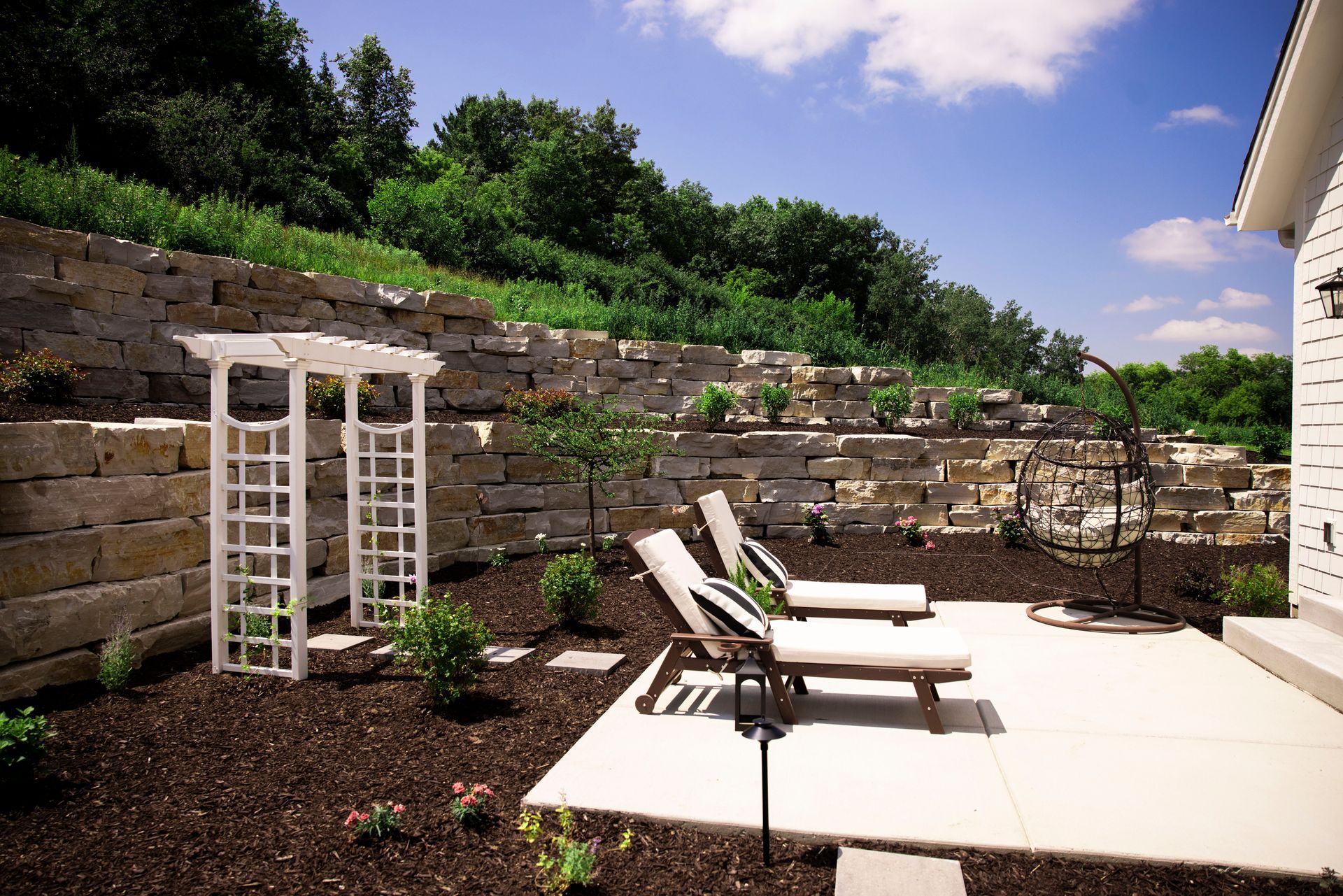 Patio with lounge chairs, white arbor, and tiered stone retaining wall against a hillside.