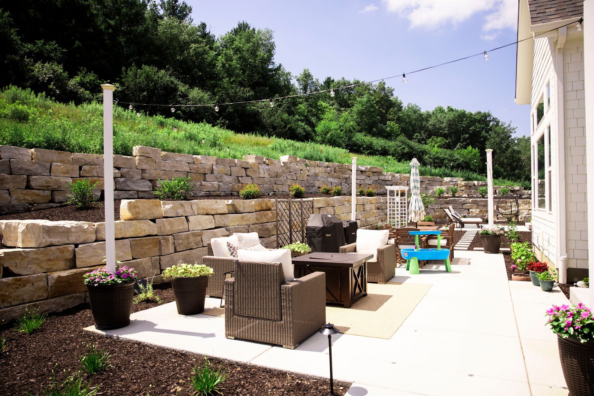 Patio with stone wall, outdoor furniture, string lights, and potted plants.