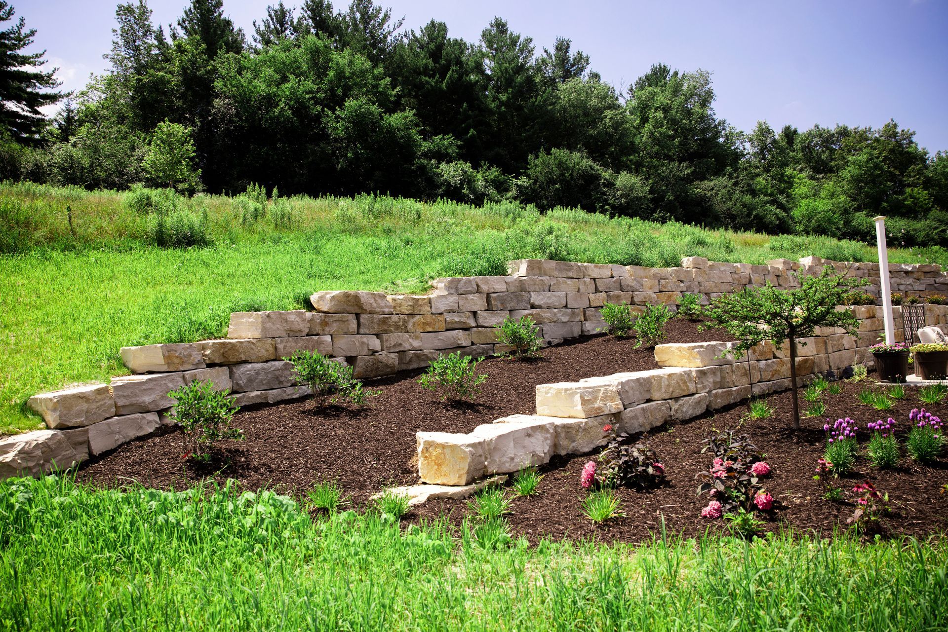 Stone retaining wall on a grassy hillside, with plants and mulch beds. Green trees in the background.